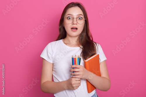 Astonished dark haired woman with opened mouth  carries textbook and colored pencils for writing or drawing  being ready to make notes while listens some shocking information  poses on pink background
