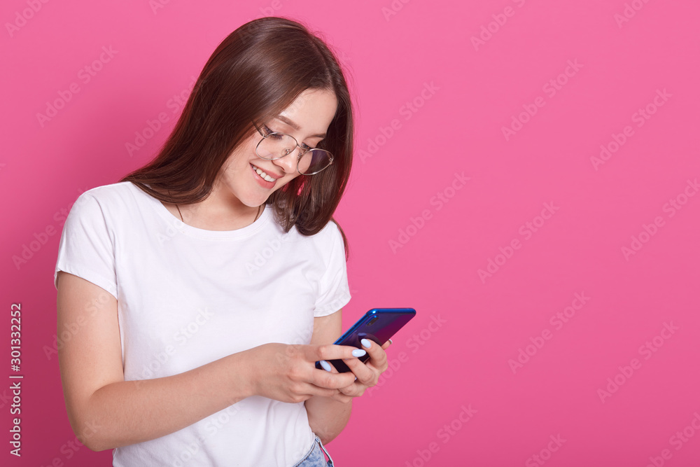 Close up portrait of adorable woman typing message to her friends or husbaund. Young female holding mobile phone and smiling while standing against pink background. Modern technology concept. Close up portrait of adorable woman typing message to her friends or husbaund. Young female holding mobile phone and smiling while standing against pink background. Modern technology concept.