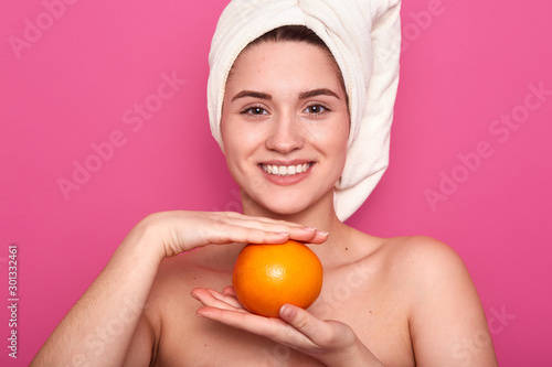 Close up portrait of young girl with nacked shoulders, holding orange, having happy facial expression. Charming female with white towel on her head, relaxing in spa salon, poses against pink wall.