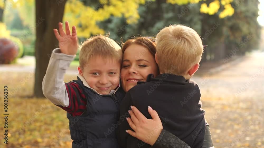 Happy young mother with little sons in autumn Park, portrait. A happy family walks in the Park in autumn, children hug their mother and wave their hands at the camera. Slow motion. Happy young mother with little sons in autumn Park, portrait. A happy family walks in the Park in autumn, children hug their mother and wave their hands at the camera. Slow motion.