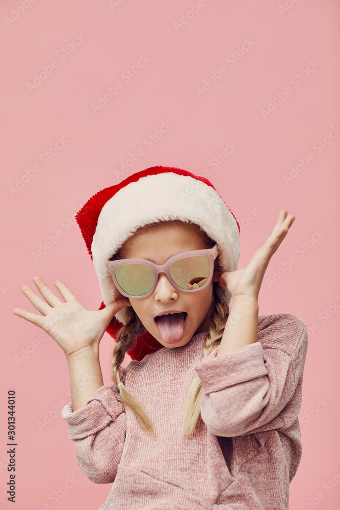 Portrait of funny little girl wearing Santa hat and fashion sunglasses posing at camera isolated on pink background Portrait of funny little girl wearing Santa hat and fashion sunglasses posing at camera isolated on pink background