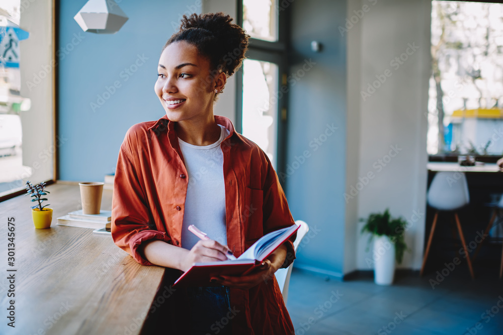 Cheerful hipster girl recalls to funny moments of day while looking away with cute smile on face during learning with notepad in cafeteria, happy dark skinned woman holding textbook for education - obrazy, fototapety, plakaty 
