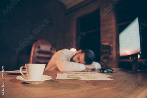 Overworked workaholic man sleep on desk feel tired on night shifts want rest at night work with white mug having hot espresso beverage diagrams standing on desktop in evening dark overnight office