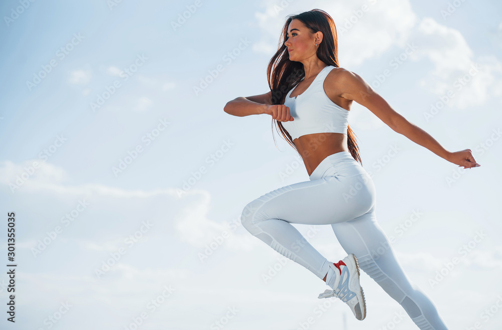 Up in the sky. Female runner in white sportive clothes doing fitness - obrazy, fototapety, plakaty 