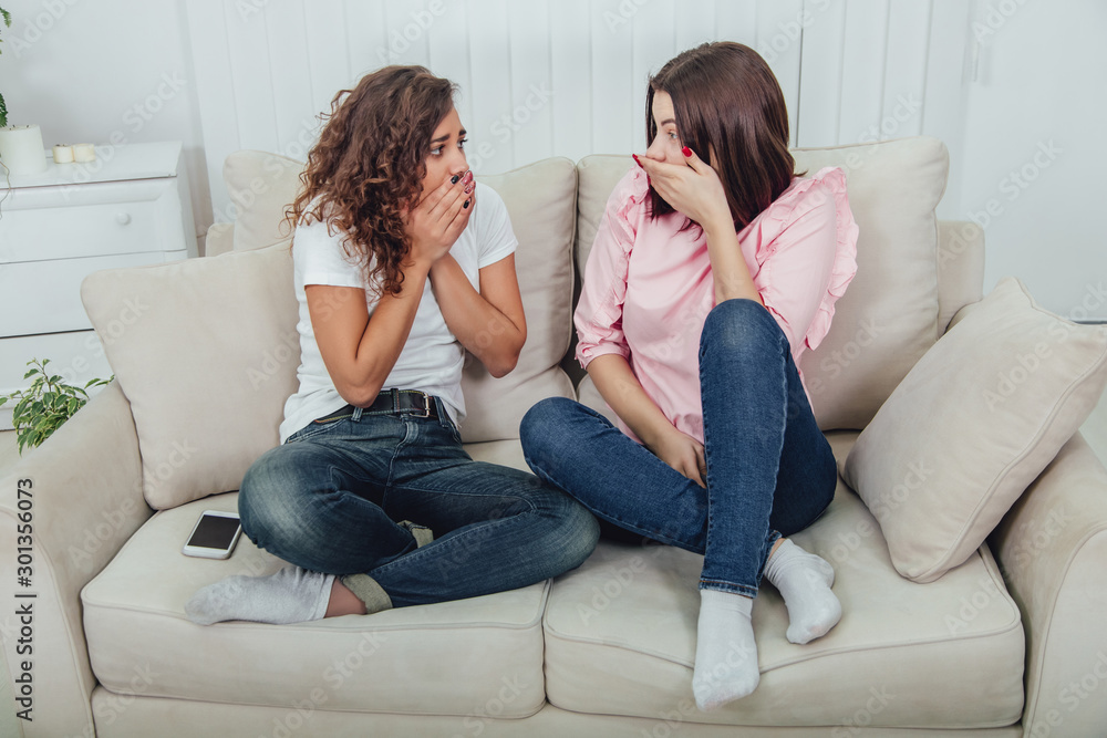 Obraz premium Two attractive girlfriends sitting on the sofa discussing something that evokes strong emotions. Surprised even scared face expressions.