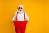 Portrait of crazy funky white bearded grandfather in santa claus hat hold big size trousers lose weight belly x-mas time show dieting effect isolated yellow color background