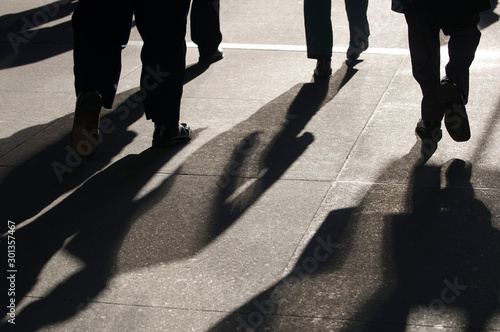 Morning sun casting dramatic shadows on the sidewalk behind silhouettes of commuting business people rushing to work © PeskyMonkey
