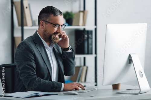 handsome businessman in formal wear talking on smartphone and looking at computer