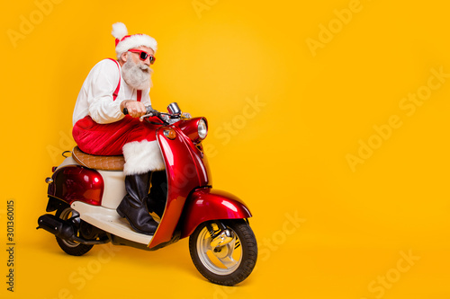 Full body photo of santa white hair grandpa rushing newyear party by bike dislike cold temperature wear trendy sun specs red pants cap shirt boots isolated yellow color background