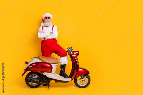 Full length photo of santa arrogant man standing self-confident near retro moped bike crossed arms wear trendy sun specs pants cap shirt isolated yellow color background
