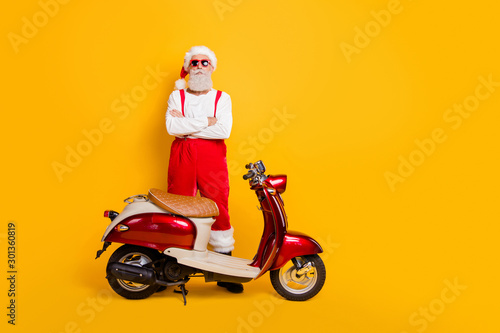 Full body photo of santa man standing confidently near retro bike crossed arms not smiling wear trendy sun specs pants cap shirt isolated yellow color background