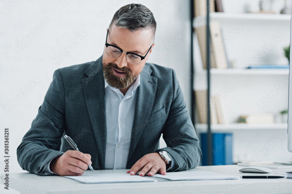 handsome businessman in formal wear and glasses writing with pen in office handsome businessman in formal wear and glasses writing with pen in office