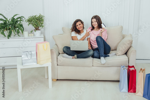Two happy female friends using laptop for shopping online. One girl is holding credit card, another pointing her finger at it.