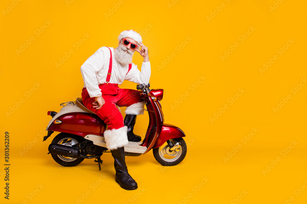 Full body photo of funny santa aged man in festive mood ready for x-mas theme party sitting on vintage bike wear sun specs pants cap shirt boots isolated yellow color background Full body photo of funny santa aged man in festive mood ready for x-mas theme party sitting on vintage bike wear sun specs pants cap shirt boots isolated yellow color background