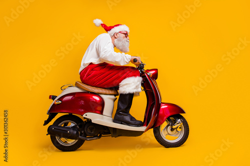Full length low angle view photo of santa white hair grandpa rushing x-mas party by moped wear sun specs costume trousers cap shirt boots isolated yellow color background
