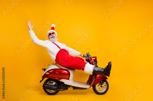 Full body photo of funny white hair santa in festive mood riding x-mas theme party by bike wear stylish sun specs trousers cap shirt boots isolated yellow color background