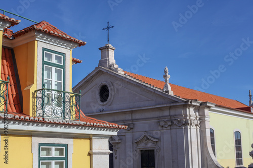 The beautiful facade of a richly finished yellow house on the large glass doors and windows, the detailed iron balconies next to a colonial-style mud-roofed church in the city of Lisbon, Portugal.