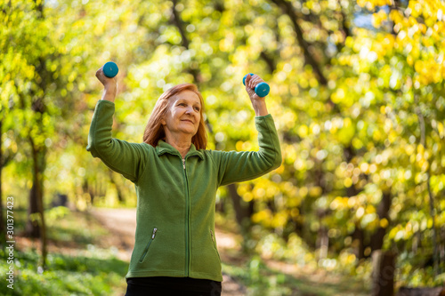 Senior woman is exercising with weights in park on sunny day.