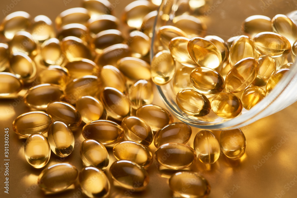 close up view of shiny fish oil capsules scattered from glass bowl on golden background close up view of shiny fish oil capsules scattered from glass bowl on golden background