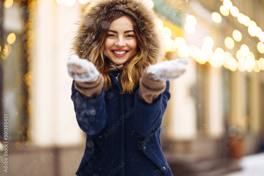 Beautiful  girl in a blue jacket and knitted hat and mittens posing in street of city. Garland lights. Enjoying snow, smiling to camera, cheerful  new year mood, true emotions. Winter fashion. - obrazy, fototapety, plakaty 