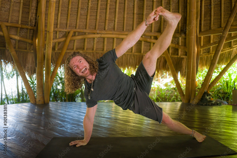lifestyle portrait of young attractive and happy man in hipster yogi style doing yoga drill on mat at beautiful Asian bamboo hut stretching in mind balance and good vibes