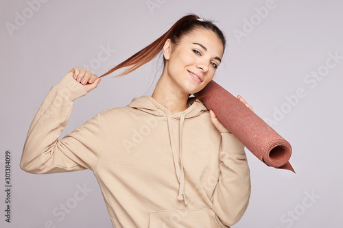 Portrait of cheerful fitness woman keeps hand on waist, rolled yoga mat, being in good physical shape, wears beige hoodie, turns gaze away with happy smile, enjoys  time in gym, motivates to sport.