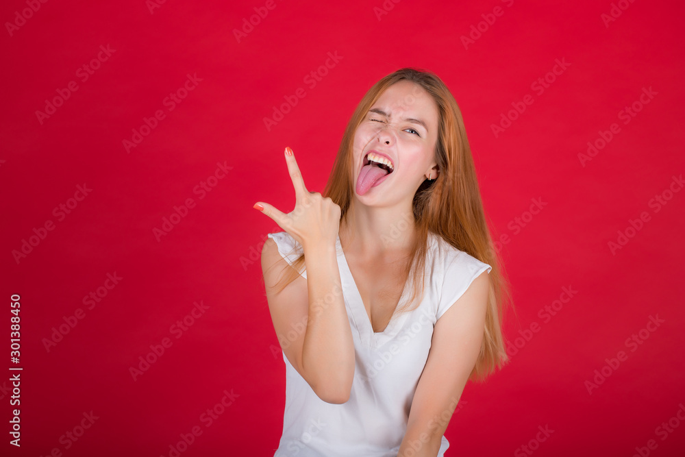 Portrait of a nice attractive cheerful red-haired girl in a white blouse shows tongue and two fingers on her right hand isolated on a bright red background Portrait of a nice attractive cheerful red-haired girl in a white blouse shows tongue and two fingers on her right hand isolated on a bright red background