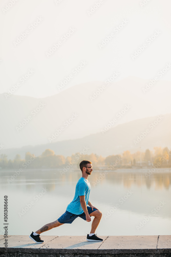 Portrait of a young man in sportswear working out by the lake in a city park. Runner warming up in the park by the water at sunset.