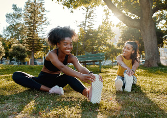 Young diverse female friends sitting on green grass stretching her legs in the morning sunlight at park - diverse friends warming up before doing group exercise 
