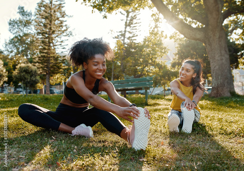 Young diverse female friends sitting on green grass stretching her legs in the morning sunlight at park - diverse friends warming up before doing group exercise  photo