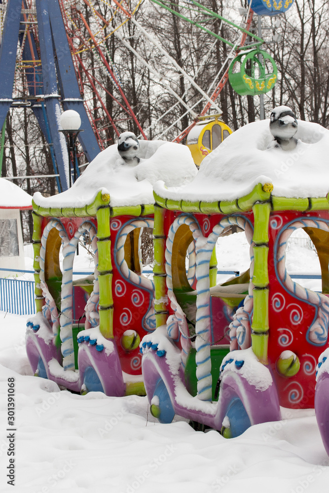 An empty amusement park in winter, a multi-colored carousel steam engine under a thick layer of snow. City leisure park for children