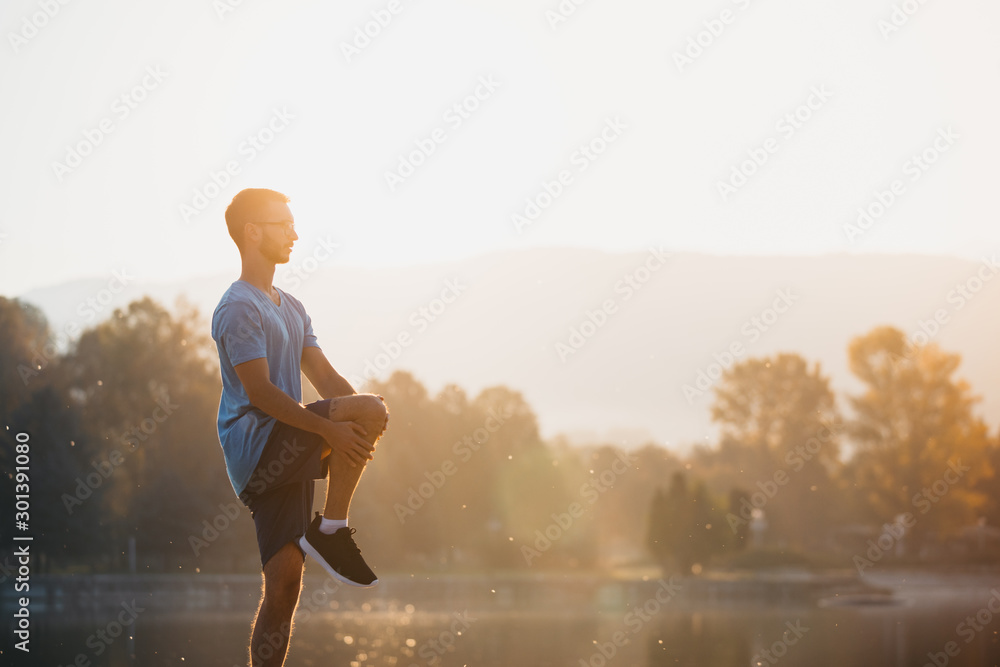 Young man in sportswear stretching by the lake in a city park. Runner warming up in the park by the water in the early morning. - obrazy, fototapety, plakaty 