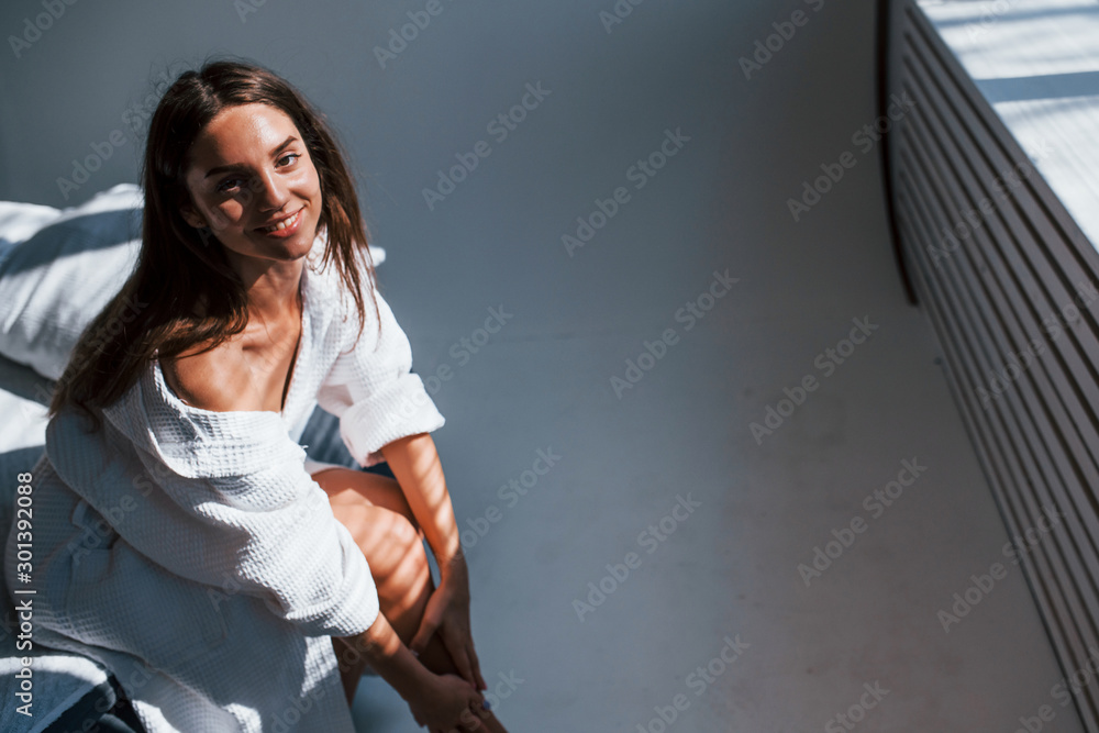 Portrait of beautiful brunette in white clothes that sits on the bed in room. Nice lighting Portrait of beautiful brunette in white clothes that sits on the bed in room. Nice lighting