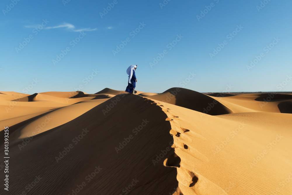 Traditional dressed Moroccan man with turban stands on a sand dune in the Sahara desert.