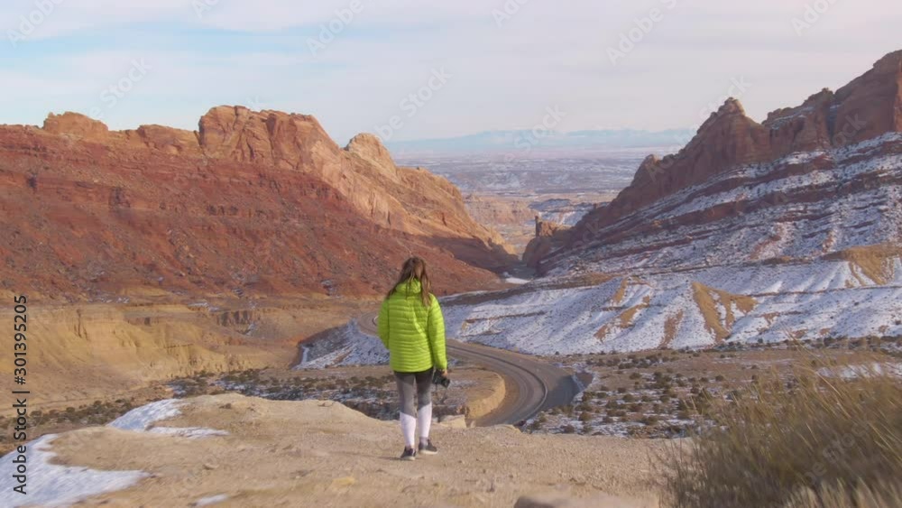 DRONE: Female traveler walks with her camera in hand along a scenic trail overlooking an asphalt highway crossing the canyon. Instagrammer walking to a good photo spot in a stunning geological park. DRONE: Female traveler walks with her camera in hand along a scenic trail overlooking an asphalt highway crossing the canyon. Instagrammer walking to a good photo spot in a stunning geological park.