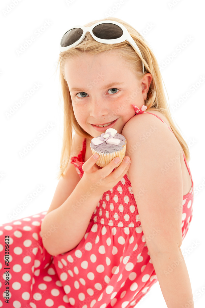 Beautiful young blonde girl wearing a pink and white polka dot dress with white framed sunglasses on her head, holding up a cup cake. Isolated on white studio background