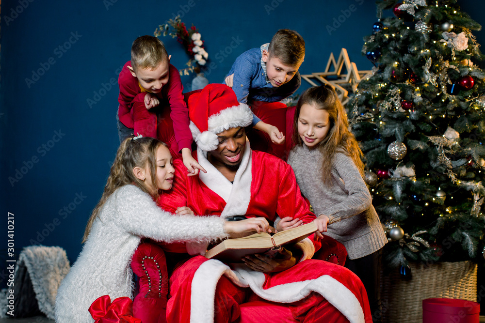 On Christmas Eve, Santa Claus reads the Christmas story for children who listen to him from all sides, on the background of a Christmas tree.