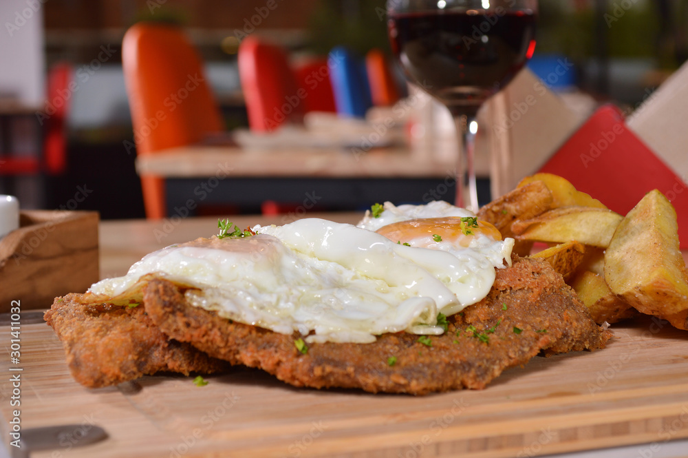 CLASSIC BREADED FRIED MEAT , KNOWN IN ARGENTINA AS MILANESA, TWO SUNNY SIDE UP EGGS, SERVED WITH FRENCH FRIES ON A WOODEN TABLE