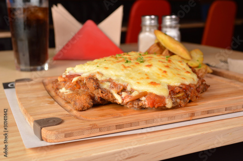 CLASSIC BREADED FRIED MEAT , KNOWN IN ARGENTINA AS MILANESA, CHEESE AND TOMATO, SERVED WITH FRENCH FRIES ON A WOODEN TABLE