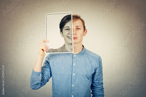 False portrait of a sad boy teenager hiding half face using a photo print paper sheet with a fake happy emotion. Mask for hiding the real expression, create new fake identity, grey wall background. photo