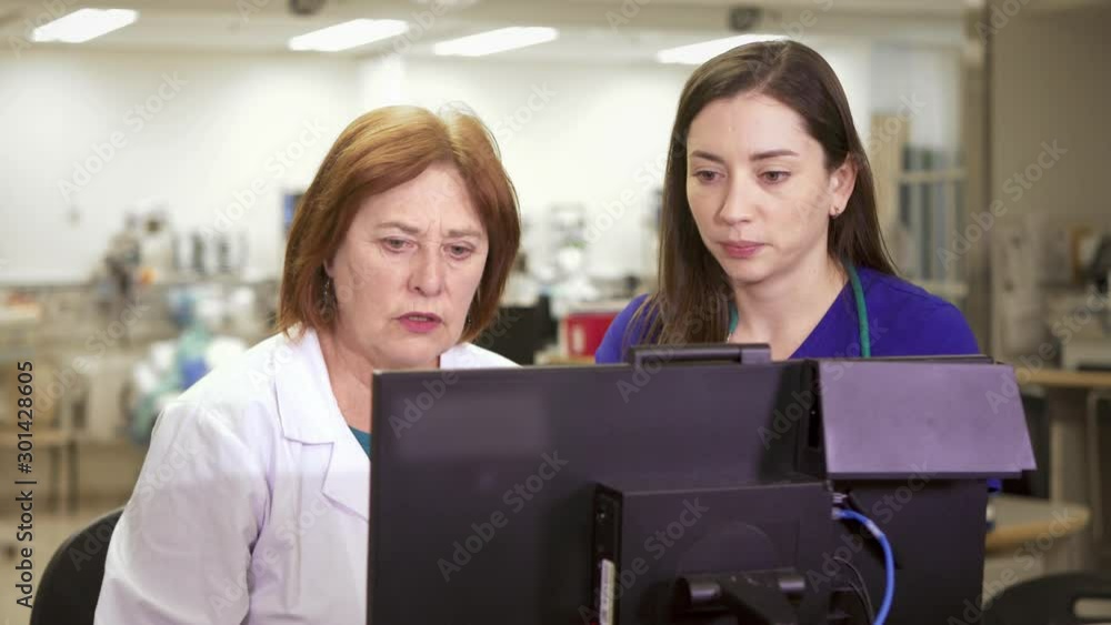A female doctor and nurse collaborate over a hospital computer. Multigenerational teamwork in a medical environment. A female doctor and nurse collaborate over a hospital computer. Multigenerational teamwork in a medical environment.