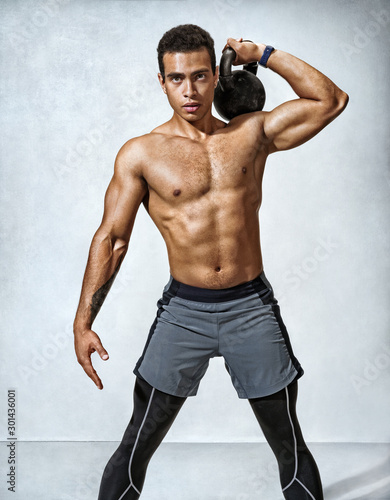 Sporty young man working out with a kettlebell. Photo of muscular man with perfect physique on grey background. Strength and motivation