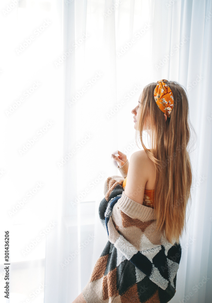 Fototapeta premium Beautiful sensual young woman fair long hair girl wearing in cozy knitted cardigan near window at the home