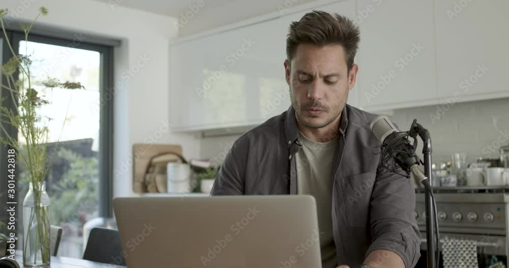 Adult male recording podcast on laptop at home in kitchen Adult male recording podcast on laptop at home in kitchen