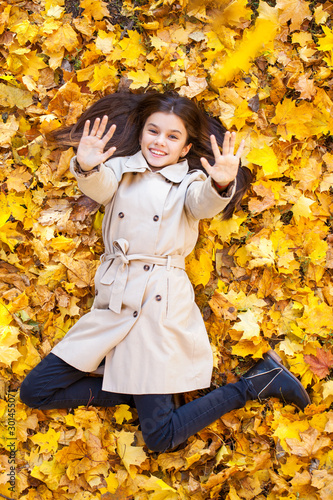 Young beautiful little girl in beige coat lying on yellow leaves