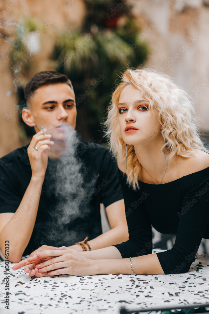 Fototapeta premium Amazing guy with a cigarette in his hand looks at a blond girl in a black dress, standing with her back bent and putting her elbows on the table.