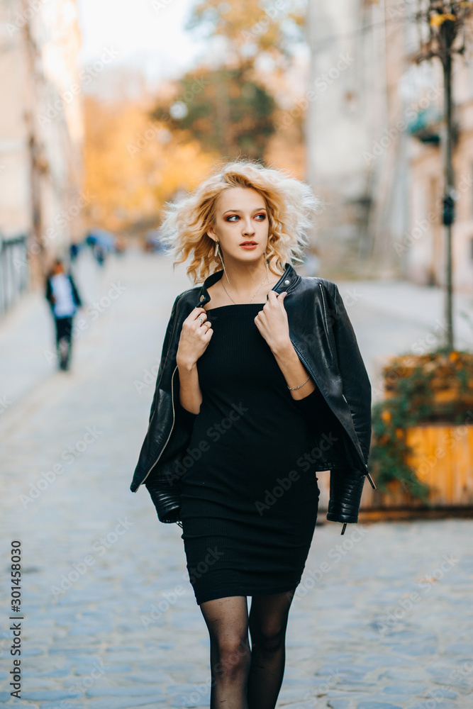 Luxury blonde in elegant evening black dress and leather jacket walks down the street