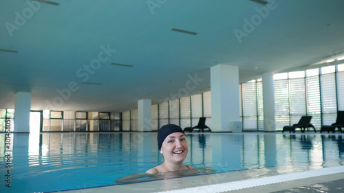 Happy woman in cap and swimsuit in swimming pool looks at camera, copy space
