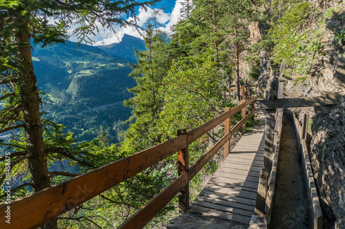 Fototapeta Naklejka Na Ścianę i Meble -  Brown wooden path with railing on the side of a mountain surrounded by trees with a valley in the background and mountains with snow on the other side.
