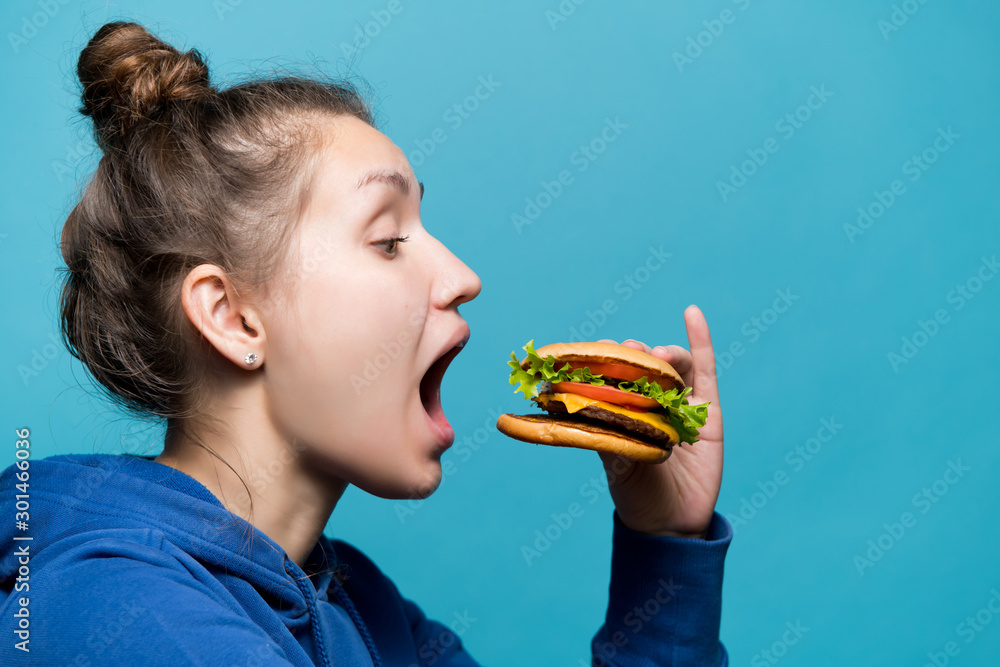 a girl in a sports sweatshirt is about to bite off a piece of a burger and opened her mouth wide at him, side view a girl in a sports sweatshirt is about to bite off a piece of a burger and opened her mouth wide at him, side view
